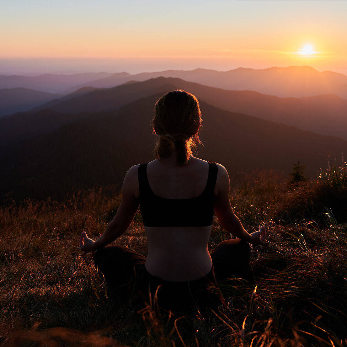 Meditating female is relaxing in mountains. Back view of woman practicing yoga in Lotus pose in the mountains. Meditating female is relaxing and watching sunset. Concept of healthy lifestyle.